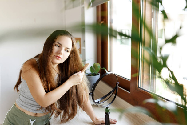 Happy young woman combing her hair and smiling looking at the mirror. Everyday hair routine concept - Photo, Image
