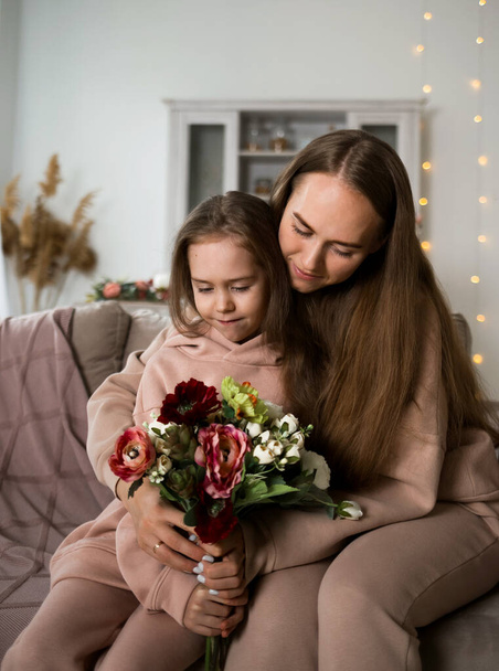 piękna mama i córka w bluzach siedzą na kanapie i świętują Dzień Matki z kwiatami - Zdjęcie, obraz