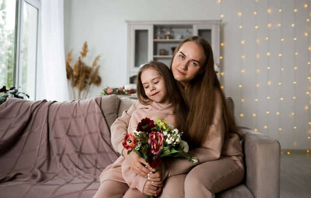 beautiful mother and daughter in sweatshirts sitting on a sofa with flowers - Photo, Image