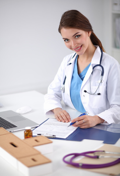 Hermosa joven sonriente doctora sentada en el escritorio y escribiendo. - Foto, Imagen