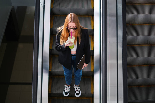 bela jovem caminhando na escada rolante de um shopping center com café em suas mãos - Foto, Imagem