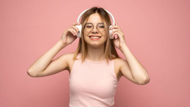 Beautiful attractive young blond woman wearing pink t-shirt and glasses in white headphones listening music and smiling on pink background in studio. Relaxing and enjoying. Lifestyle. - Fotografie, Obrázek