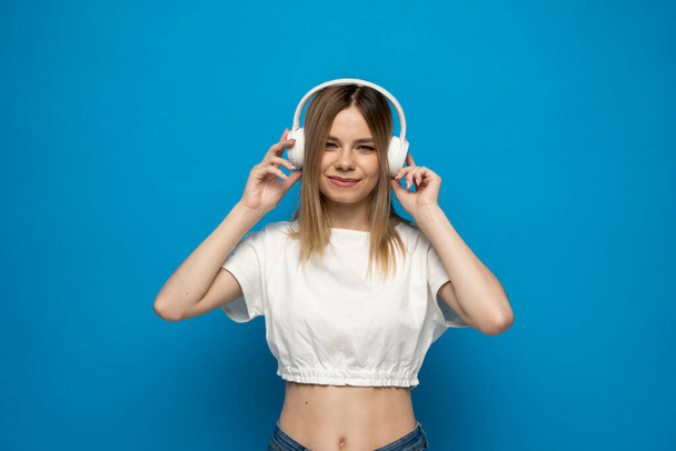 Beautiful attractive young blond woman wearing white t-shirt and glasses in white headphones listening music and dancing on blue background in studio. Relaxing and enjoying. Lifestyle. - Valokuva, kuva