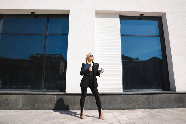 A young woman in a stylish suit with a laptop and a cup of coffee poses against the background of a business center - Foto, immagini