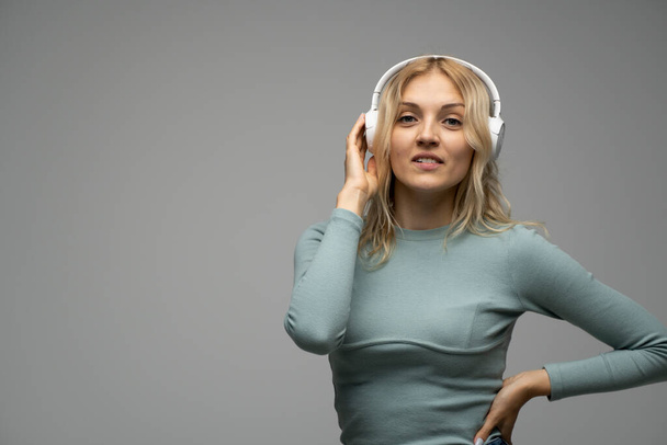 Beautiful attractive young blond woman wearing blue t-shirt and glasses in white headphones listening music and smiling on grey background in studio. Relaxing and enjoying. Lifestyle. - Φωτογραφία, εικόνα