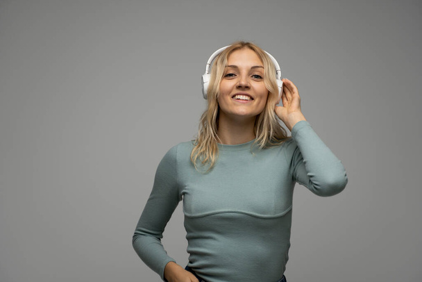 Beautiful attractive young blond woman wearing blue t-shirt and glasses in white headphones listening music and smiling on grey background in studio. Relaxing and enjoying. Lifestyle. - Fotó, kép