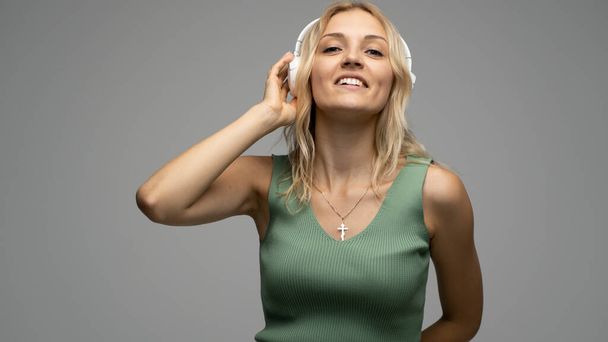 Beautiful attractive young blond woman wearing green t-shirt and glasses in white headphones listening music and smiling on grey background in studio. Relaxing and enjoying. Lifestyle. - Photo, Image