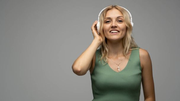 Beautiful attractive young blond woman wearing green t-shirt and glasses in white headphones listening music and smiling on grey background in studio. Relaxing and enjoying. Lifestyle. - Foto, afbeelding