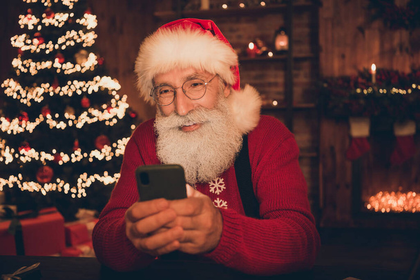 Photo of cheerful santa claus look take remote phone order wear hat pullover in north pole office indoors - Фото, зображення