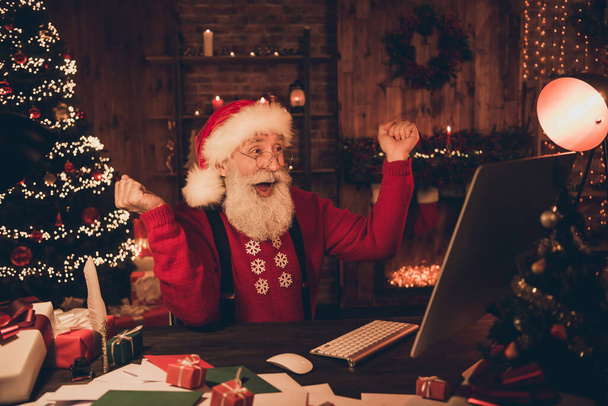 Photo of astonished santa claus look pc screen raise fists wear hat pullover in north pole office indoors - Foto, afbeelding