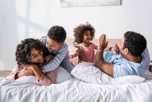 幸せなアフリカ系アメリカ人男の子抱擁お母さん近くお父さんと妹遊びパティケーキゲームでベッド  - 写真・画像