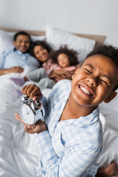 excited african american boy with closed eyes holding alarm clock near blurred family in bed - Foto, Imagem