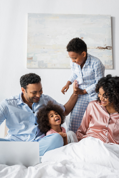 african american man smiling near laptop and happy family on bed - Photo, Image