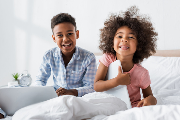 aufgeregte afrikanisch-amerikanische Kinder schauen in die Kamera, während sie mit Gadgets im Bett sitzen - Foto, Bild