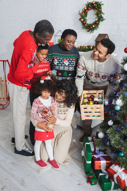 щасливий афроамериканський чоловік тримає дерев'яну коробку з різдвяними кульками поблизу сім'ї
  - Фото, зображення