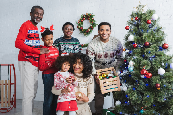 man holding wooden box with decorative baubles near christmas tree and african american family - Photo, Image