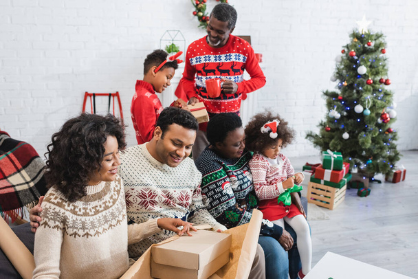 χαρούμενη αφρικάνικη αμερικανική οικογένεια συσκευασίας δώρα στο σαλόνι με θολή χριστουγεννιάτικο δέντρο - Φωτογραφία, εικόνα