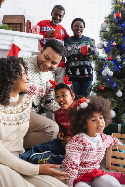 cheerful african american family near decorated christmas tree and blurred grandparents with cups - Valokuva, kuva
