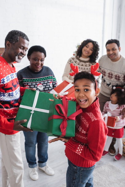 amazed african american boy in headband with reindeer horns showing christmas present near blurred family - Photo, Image