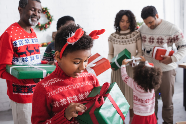amazed african american boy in reindeer horns headband holding xmas present near blurred family with gift boxes - Photo, Image