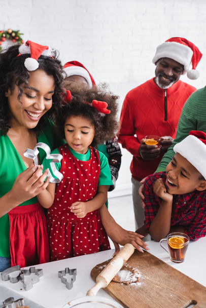 african american woman showing cookie cutter to daughter near dough and happy family with orange cinnamon tea - Photo, Image
