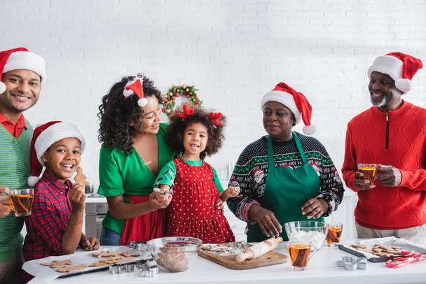 happy girl holding cookie cutter while preparing christmas cookies with african american family - 写真・画像