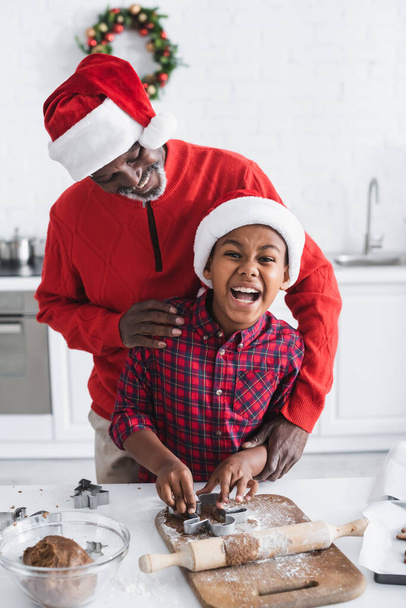 excited african american boy in santa hat cutting dough with cookie cutter near happy grandpa in kitchen - Foto, imagen