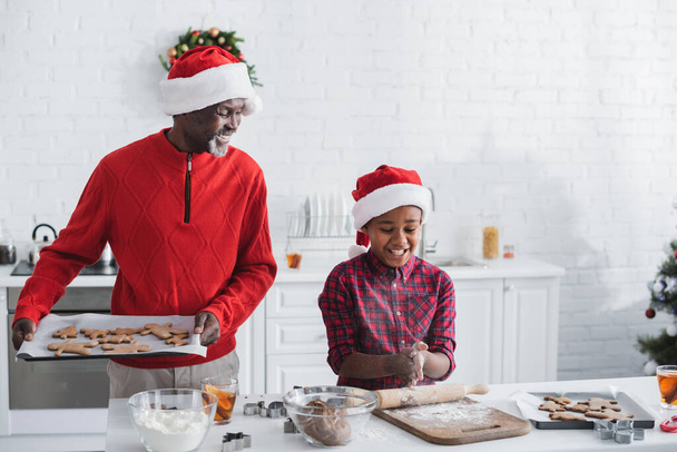 hombre afroamericano feliz sosteniendo bandeja para hornear con galletas de Navidad cerca de nieto  - Foto, Imagen