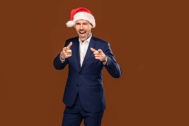 Happy handsome businessman wearing santa claus cap pointing his fingers to the camera on brown studio background and smiling. - Фото, зображення
