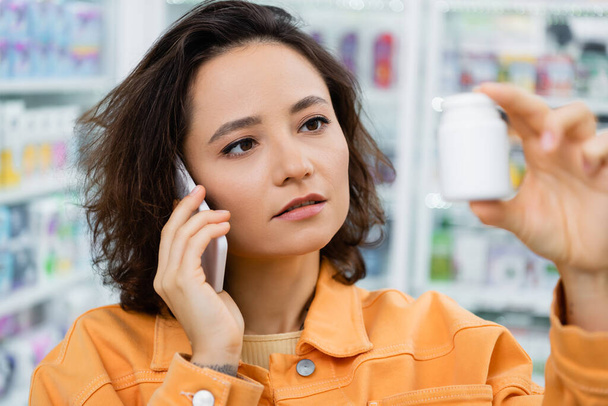 brunette woman holding bottle with medication while talking on smartphone in drugstore - Фото, изображение