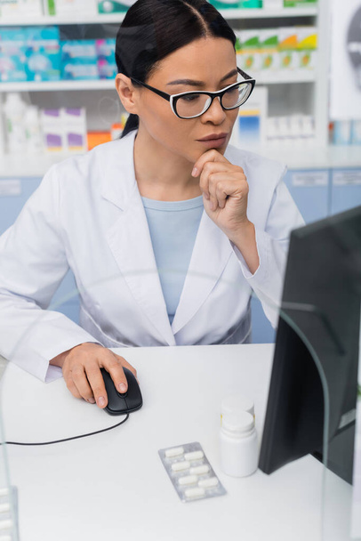 brunette asian pharmacist in glasses and white coat using computer in drugstore - Photo, Image