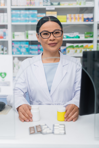 wesoły azjatycki farmaceuta trzymający butelki z lekami w pobliżu blistrów na biurku przy ladzie - Zdjęcie, obraz