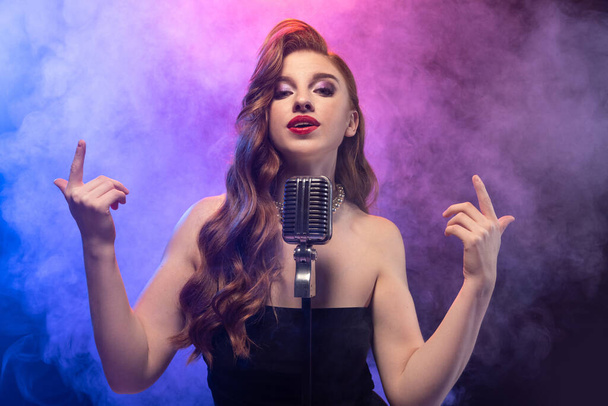 Retrato recortado de hermosa cantante femenina expresiva en movimiento aislado sobre el fondo azul púrpura degradado en luz de neón con humo de escenario - Foto, imagen