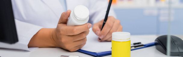 обрезанный вид аптекаря, держащего бутылку с лекарствами и пишущего на планшете, баннер - Фото, изображение