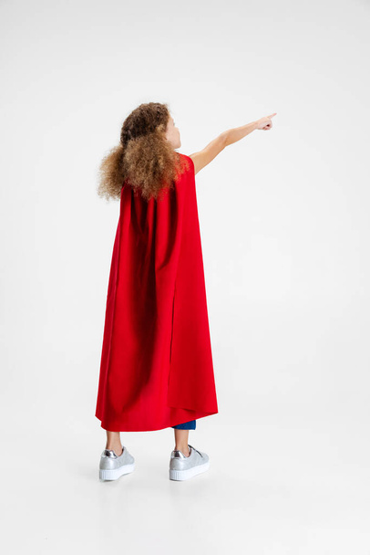 Back view of little girl, kid standing at her back to camera wearing red cape isolated on gray studio background with copyspace for ad - Fotografie, Obrázek
