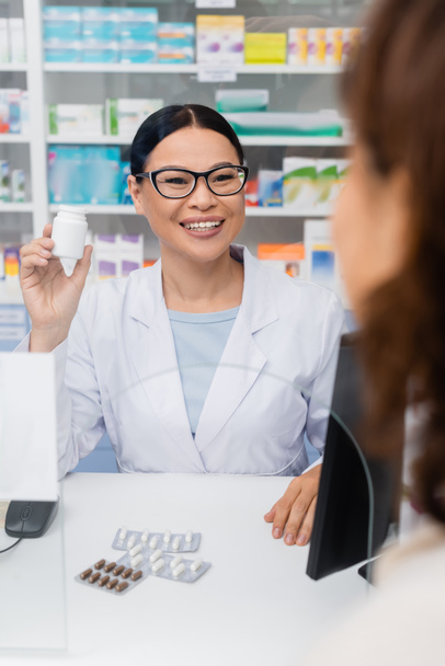 cheerful asian pharmacist holding bottle with medication near blurred customer - Foto, afbeelding