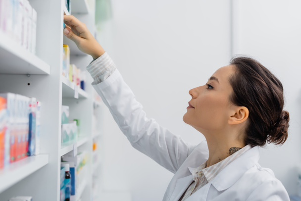 tattooed apothecary in white coat reaching shelf with medication in drugstore - Photo, Image