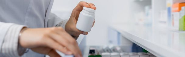 vista recortada del farmacéutico en la botella de la celebración de la capa blanca con la medicación mientras que abre el cajón en la farmacia, bandera - Foto, imagen