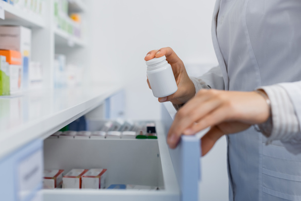 cropped view of pharmacist in white coat holding bottle with medication while opening drawer in drugstore - Foto, immagini