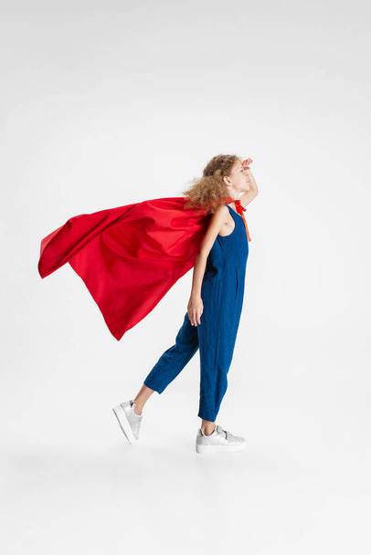 Portrait of beautiful caucasian little curly girl wearing red cape standing, walking isolated on gray studio background with copyspace for ad - Fotografie, Obrázek