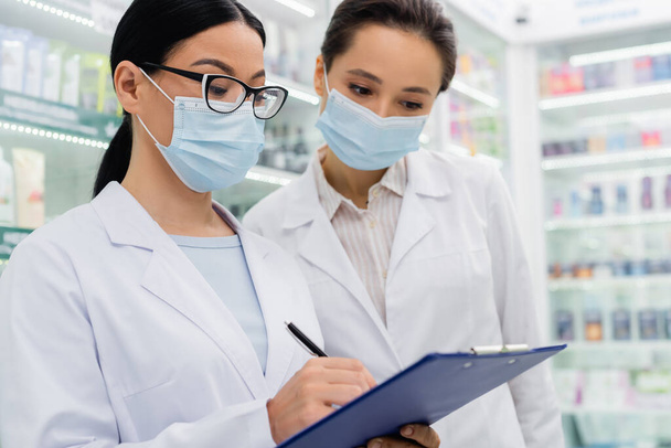 asian pharmacist in medical mask writing on clipboard near colleague  - Photo, Image