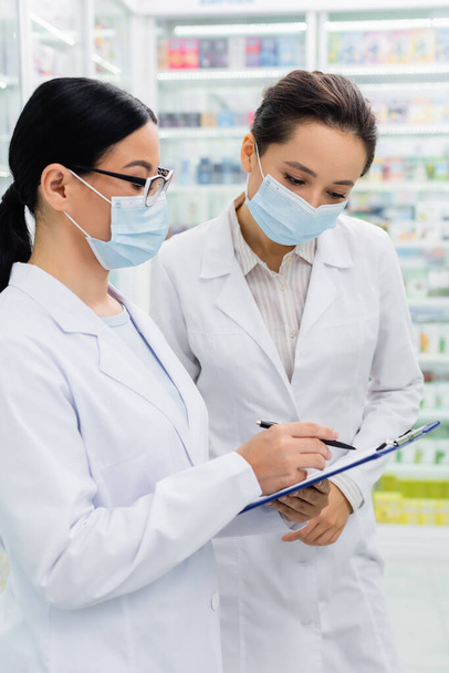 asian pharmacist in glasses and medical mask writing on clipboard near colleague  - Foto, afbeelding