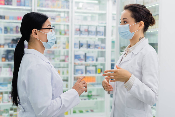 farmacisti interrazziale in maschere mediche che si guardano mentre parlano in farmacia - Foto, immagini
