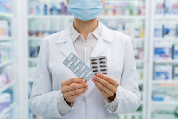 oříznutý pohled na lékárníka v lékařské masce držící blistry s léky  - Fotografie, Obrázek