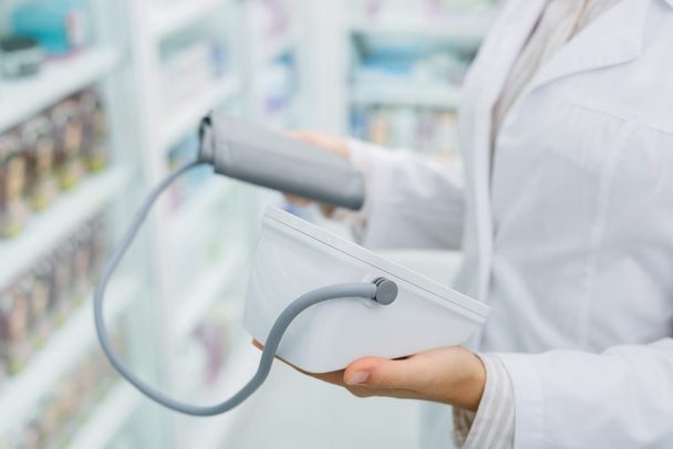 oříznutý pohled lékárníka v bílém plášti držícího digitální tonometr v lékárně - Fotografie, Obrázek