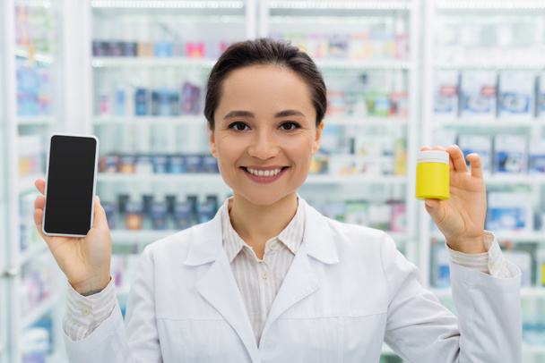 cheerful pharmacist in white coat holding smartphone with blank screen and bottle in drugstore - Фото, изображение
