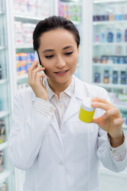 cheerful pharmacist in white coat talking on smartphone and holding bottle in drugstore - Foto, afbeelding