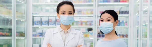 pharmacist in medical mask standing near asian customer in drugstore, banner - 写真・画像