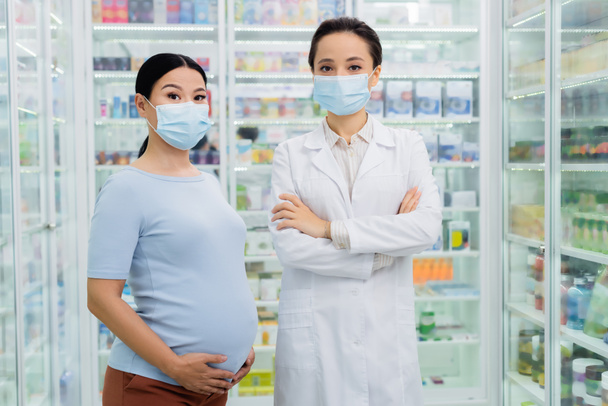 pharmacist in medical mask standing with crossed arms near asian pregnant woman in drugstore  - Photo, Image