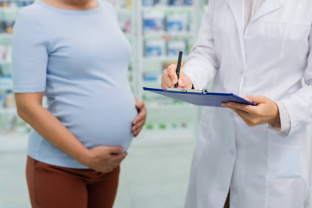 cropped view of pregnant woman standing near pharmacist writing on clipboard - Foto, afbeelding